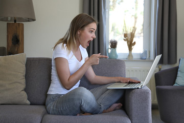 Hübsche blonde Frau sitzt auf einer Couch und zeigt begeistert auf einen Laptop