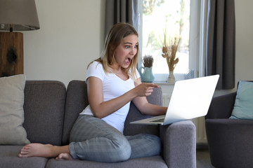 Obraz premium Hünsche blonde Frau sitzt auf einem Sofa und zeigt begeistert auf einen Laptop