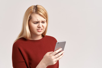 Naklejka premium Worried upset nervous female model bites thumb, holds modern smart phone, feels frustrated as has failure of payment. Disappointed woman reads message about postponing or canceling interview