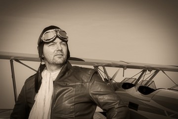 Portrait of a vintage pilot with leather cap, scarf and aviator glasses in front of a historic airplane biplane - Portrait of a man in historical pilot clothing - vintage old picture style
