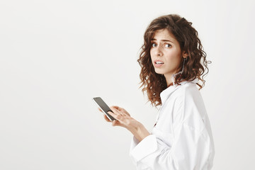 Profile indoor portrait of confused and clueless caucasian woman holding smartphone and looking at camera with frowned eyebrows and dissatisfied look, being unhappy of message she received