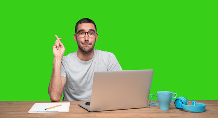 Hopeful young man sitting at his desk with crossed fingers gesture - Green background
