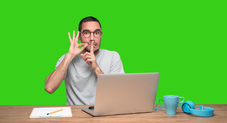 Confident young man sitting at his desk with a gesture of keeping a secret