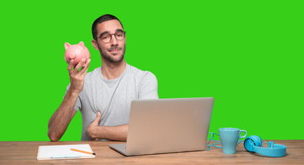 Happy young man sitting at his desk and holding a piggy bank - Green background