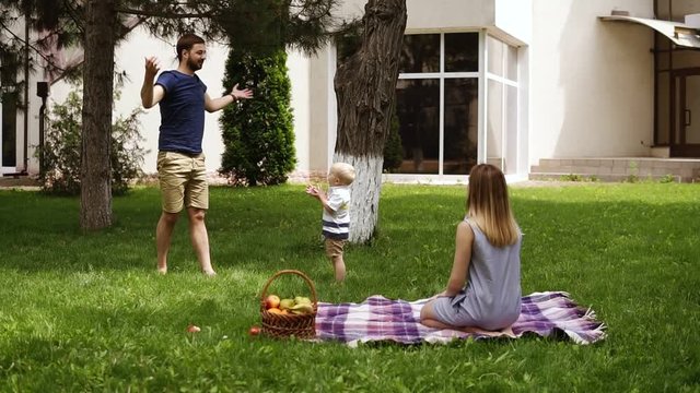 Front view of an idyll family hanging out together in the park. Young mom sitting on a grass and watching his son playing. Father amuses son, throws cones into the air. Slow motion