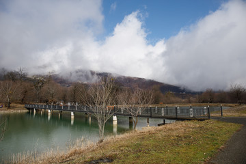 Fototapeta na wymiar Forest lake with bridge during the sunny day with winter trees and blue cloudy sky. Beautiful natural mountain lake with forest in the background and stormy clouds on the sky.
