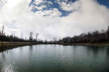 Forest lake with bridge during the sunny day with winter trees and blue cloudy sky. Beautiful natural mountain lake with forest in the background and stormy clouds on the sky.