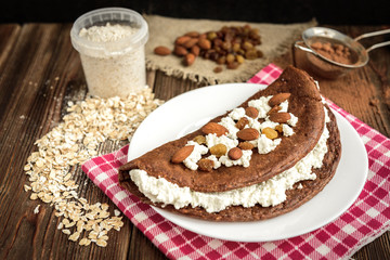 Homemade chocolate oat pancake with cottage cheese on white plate on dark wooden table.
