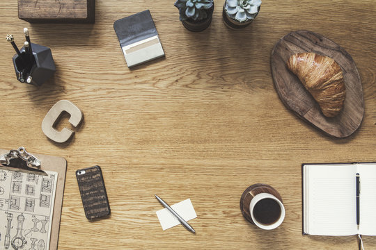 Stylish composition of flat lay on wooden desk with croissant, cup of coffee, photos, notebook, succulents and office accessories. Creative desk   of traveler with copy space.