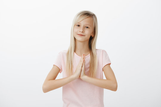 Cute obedient girl like angel for parents. Portrait of beautiful young european woman with fair hair, holding hands in pray and smiling, asking mom to buy sweets, beginning or hoping over gray wall