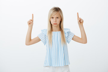 Fototapeta na wymiar Portrait of offended angry caucasian female child with long blond hair, pouting and sulking, raising index fingers and pointing up, seeing something disappointing and insulting over gray wall
