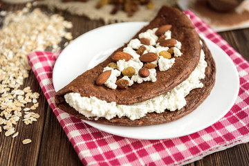 Homemade chocolate oat pancake with cottage cheese on white plate on dark wooden table.