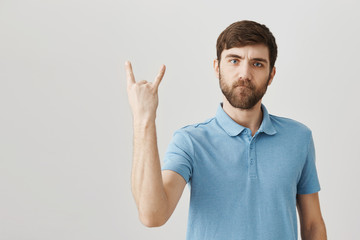 Fototapeta premium Rock n roll is alive. Portrait of serious cool european bearded man showing rock gesture with raised hand, expressing confidence and firm attitude to attend concert of favorite band