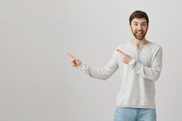 Happy good-looking caucasian bearded guy pointing left with both index fingers, smiling and being excited while standing over gray background. Upbeat boyfriend asks permission to ride roller coaster