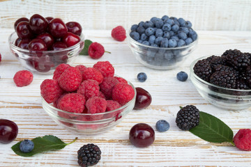 Raspberries, blackberries, cherries and blueberries in a glass bowls on wooden background. Seasonal fruits