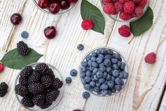 Raspberries, blackberries, cherries and blueberries in a glass bowls on wooden background. Seasonal fruits