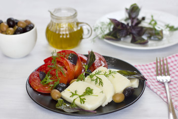 White cheese with tomatoes, green and black olives, basil, coriander and olive oil on a black plate