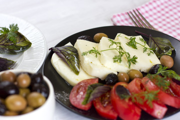 White cheese with tomatoes, green and black olives, basil, coriander and olive oil on a black plate