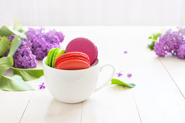 White cup with sweet colorful french macaroons, spring lilac flowers on white wooden table. Beautiful dessert Colorful almond cookies Pastel colors Bouquet of lilacs