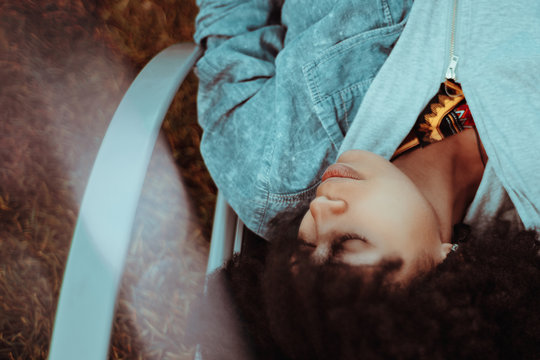 Charming adult black girl is sleeping outdoors on the recliner; Brazilian girl with curly Afro hair is napping on the daybed outside; thoughtful African American female on a street chaise-longue