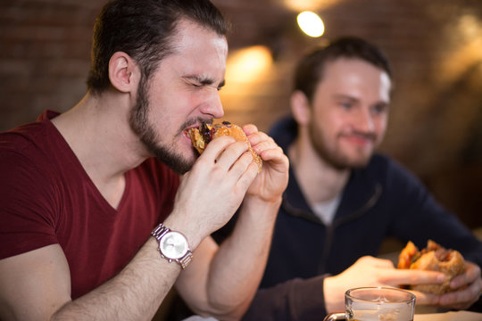 Two happy male friends sitting, talking and having lunch with burgers and beer at cozy cafe interior. One emotional guy making a good bite of tasty burger