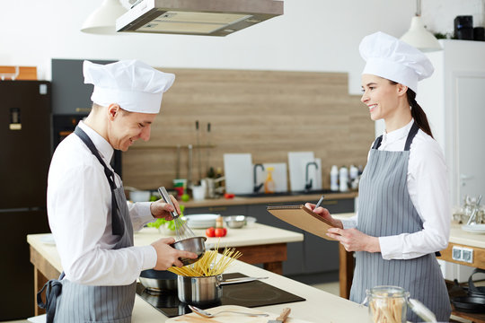 Two young trainees in chef uniform and aprons cooking together in the kitchen