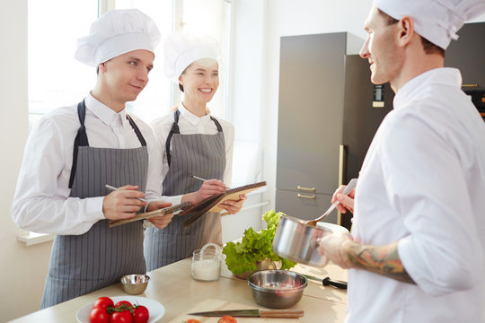 Happy young man and woman in chef uniform making notes while listening to their master in the kitchen