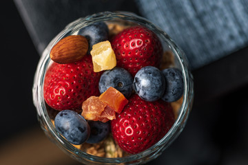 A healthy colourful snack consisting of granola and yoghurt with assorted berried and dried fruits in a small glass cup.


