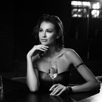 Elegant young woman sitting in bar and having good time alone. Pretty brunette girl in black leather dress. Lady drinking glass of wine.