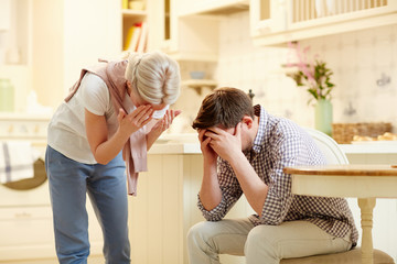 Upset young husband and wife hiding their faces in hands while grieving in the kitchen