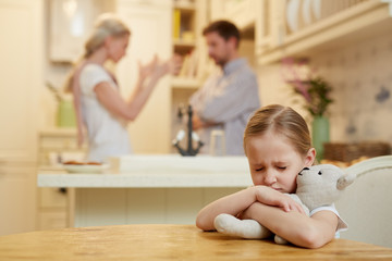 Fototapeta premium Upset little girl with teddybear sitting by table while her parents rowing on background