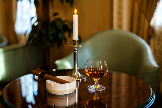 A glass of cognac, cigar and candles on a wooden table.