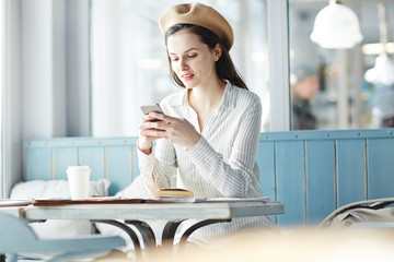 Young stylish woman with smartphone messaging while having rest in cafe
