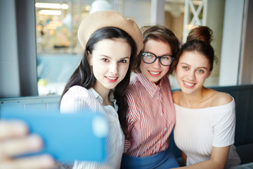 Young girlfriends relaxing in cafe and making selfie on smartphone