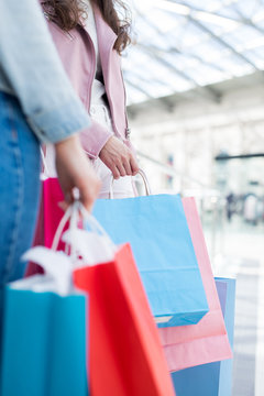 Close-up of unrecognizable girls in casual outfits holding colorful paper shopping bags while walking over mall together