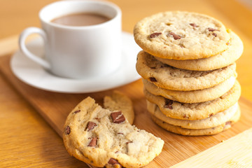 Stack of chocolate chip cookies on wooden board with white cup in the background