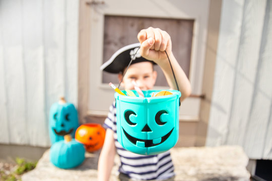 Boy In A Pirate Costume Holds A Bucket With Inedible Gifts.  Teal Pumpkin Project. Alternative Non-food Treats For Kids With Food Allergies. The Concept Of Health For Children In The Halloween Season.