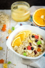 Homemade oatmeal porridge with honey, orange, raisin, peanut and candied fruit on white wooden background. Healthy breakfast.