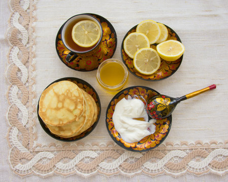 tea in a painted Cup, on a saucer sliced lemon, pancakes, sour cream and a wooden spoon for overlaying. tea party in rustic style