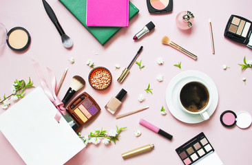 Various cosmetic products for makeup and white spring flowers in a white gift bag, notepad, cup of coffee on a pink pastel background top view with copy space