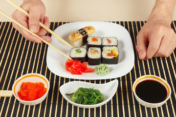 Hands taking sushi with chopsticks. Sushi set, chuka salad, soy sauce and salmon caviar on a black bamboo mat.