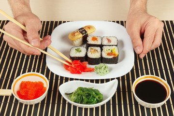 Hands taking ginger with chopsticks. Sushi set, hiyashi wakame salad, soy sauce and salmon caviar on a black bamboo mat.