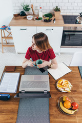 Woman holding cup sitting at table with laptop front of her in the kitchen.