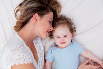 Young mother with her 7 month old little son dressed in pajamas are relaxing and playing in the bed at the weekend together, lazy morning, warm and cozy scene.