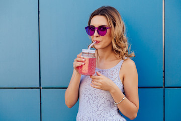 Young trendy woman in sunglasses sipping fresh cold beverage, drinking through the straw, looking at camera, outdoors.