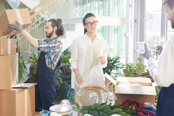 Positive confident female interior designer in glasses holding twig while talking to mover while they unpacking boxes and examining goods in space full of plants