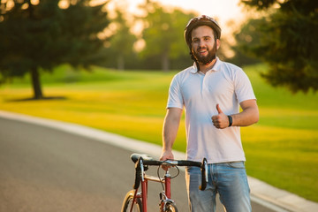 Portrait of happy handsome guy with bicycle. Cute young smiling cyclist giving thumb up outdoors. Enjoying summer weekend. Vacation on bike.