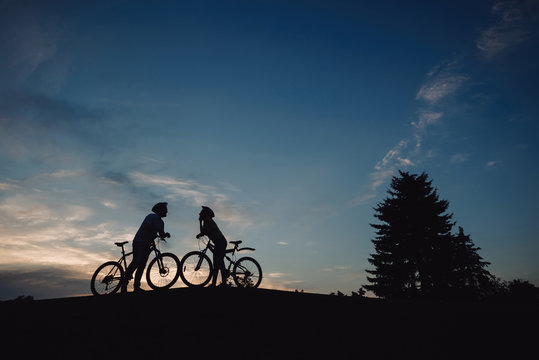 Couple of cyclists on evening sky background. Young man and woman with bikes loking ar each other at sunset sky. Enjoying the evening scenery.