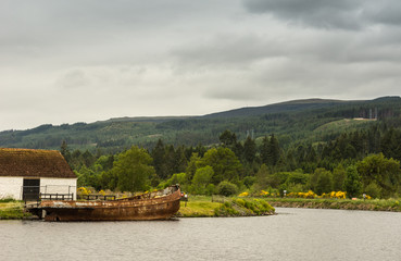 Fototapeta premium Fort Augustus, Scotland - June 11, 2012: Oich Canal behind the locks. Bucolic scenery with rusted boat wreck. Green hils with yellow flowers on horizon under heavy gray sky.