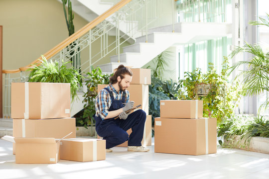 Serious Concentrated Handsome Young Male Moving Company Manager In Comfortable Uniform Crouching While Checking Packaged Boxes And Writing Out Information From Labels In Lobby